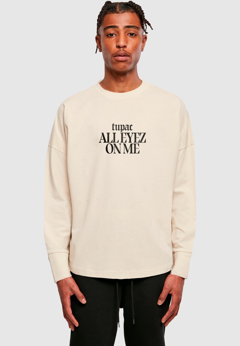 Sweatshirt bej cu mânecă lungă și text negru care spune "tupac ALLEYEZ ON ME", având un guler tip crew și o croială relaxată. Pantaloni negri vizibili.