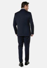 Marineblauw pak met een getailleerde pasvorm; inclusief een enkele split, slanke revers en bijpassende broek. Gedragen met zwarte dresschoenen.
