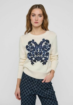 Donna che indossa un maglione crema con ricamo floreale blu scuro e pantaloni scuri a fantasia, in piedi con una mano in tasca su uno sfondo semplice.