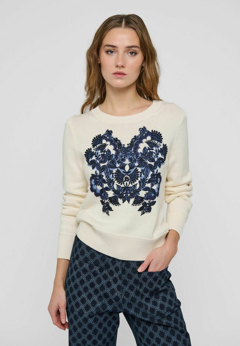 Femme portant un pull crème avec une broderie florale bleu foncé et un pantalon sombre à motifs, debout avec une main dans la poche devant un fond uni.