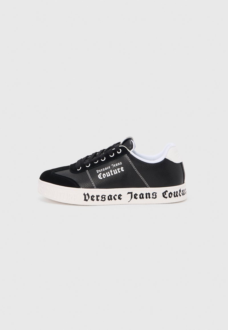 Ténis baixo preto com sola branca, apresentando o texto "Versace Jeans Couture" no lado e na sola, atacadores pretos e colarinho branco no calcanhar.