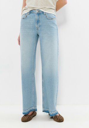 Jean bleu clair à jambe droite avec des clous argentés sur les poches avant, porté avec des chaussures plates marron cloutées, sur une personne debout.