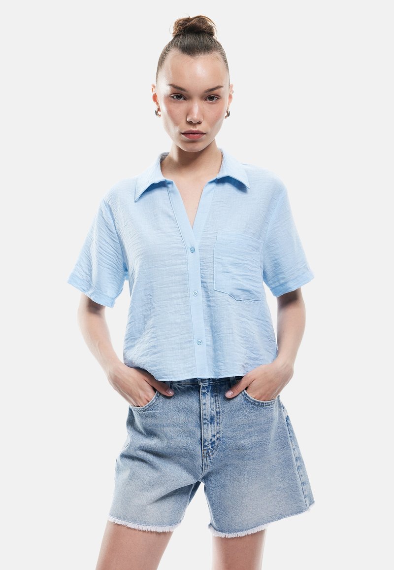 Ung kvinde med hår i en knold iført en lys blå kortærmet skjorte med knapper og højtaljede denimshorts med hænderne i lommerne.