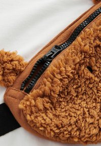Brun fleecepåse med texturerad yta och svart dragkedja, fäst vid en vit textilbakgrund. Har en mjuk, fluffig yta.