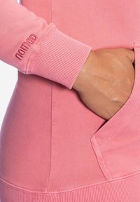 Rosa Kapuzenpulli mit gerippten Bündchen, einer Seitentasche und geprägtem Markenlogo. Der Stoff wirkt weich und strukturiert.