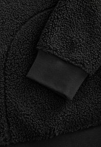 Pull en polaire noire avec une couche extérieure texturée, une doublure douce et des poignets côtelés, offrant chaleur et confort.