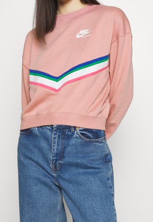 Femme portant un sweat-shirt rose Nike avec des bandes chevrons bleues, vertes, blanches et roses, associé à un jean bleu taille haute.