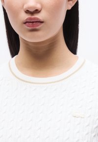 Γυναίκα με ίσια σκούρα μαλλιά φοράει λευκό πουλόβερ πλεκτό με σχέδιο καλωδίου και μικρό κεντητό λογότυπο κοντά στον γιακά.