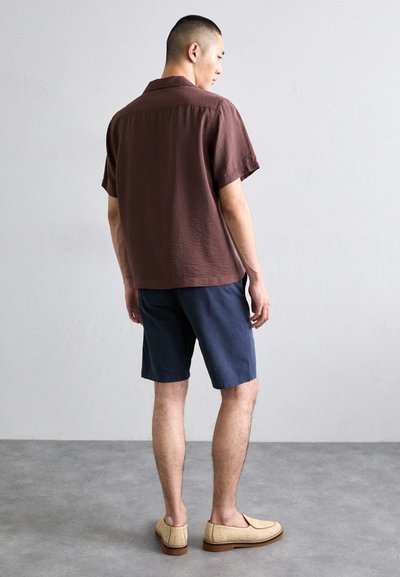 Chemise à manches courtes, texturée, de couleur marron foncé, associée à un short bleu marine. Des mocassins de couleur claire avec des détails tissés complètent l'ensemble.