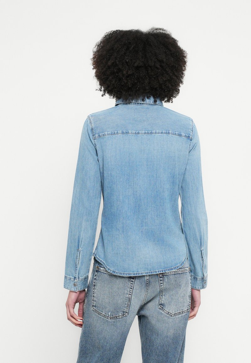 Lyseblå denimjakke med klassisk krave, lange ærmer og en buet kant. Har en glat tekstur og minimale syningdetaljer.