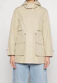 Beige huva jacka med två framfickor med klaff, insydd midja och elastiska mudd. Slät yta och lättviktig design.