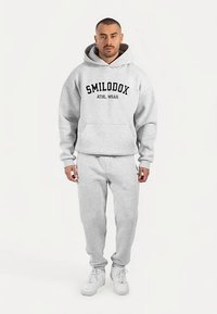 Grå hoodie med "SMILODOX ATHL. WEAR"-logotyp, känguruficka, ihop med grå mjukisbyxor och vita sneakers, med avslappnad passform.