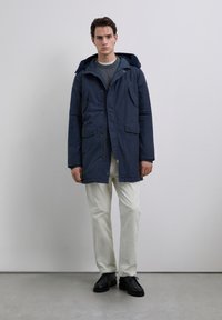 Ung mand iført navy parka med h�ætte, grå sweater, hvide bukser og sorte sko, stående foran en ensfarvet lys grå baggrund.
