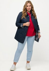 Námořnická prošívaná bunda s kostkovanou podšívkou, nošena přes červené polo tričko. Světle modré kalhoty s ohrnutými manžetami a bílé tenisky dotvářejí celý vzhled.