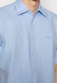 Hombre con una camisa de botones de manga corta de color azul claro, con cuello y un pequeño logotipo "BOSS" bordado en el pecho.