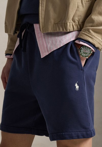 Tamsiai mėlyni medvilniniai šortai su elastinga juosmens juosta, logotipo siuvinėjimu ir šoninėmis kišenėmis. Suderinti su šviesia dryžuota palaidine ir laikrodžiu.