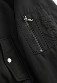 Tissu de veste synthétique noire avec une texture lisse, présentant une poche zippée et un accent en bouton-pression argenté.