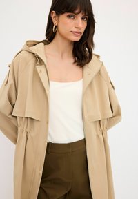 Beige huva jacka med dragkedja, elastisk midja och framlommer, bärs över en vit topp och olivgröna byxor. Guldiga detaljer.