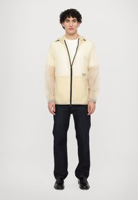 Light yellow windbreaker with a black zipper, elastic cuffs, and hood. The jacket features a logo on the left chest and a lightweight, semi-transparent design.