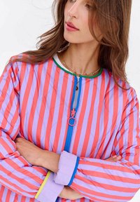 Kobieta z długimi brązowymi włosami, nosząca pikowaną kurtkę w pionowe różowe i lawendowe paski, z niebieskim zamkiem błyskawicznym oraz złożonymi rękawami z niebieskim wykończeniem.