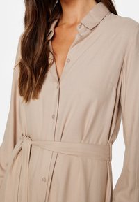 Koszula zapinana na guziki w miękkim beżowym materiale, z kołnierzykiem, długimi rękawami i związanym paskiem w talii, charakteryzująca się subtelną teksturą oraz jasnymi guzikami.