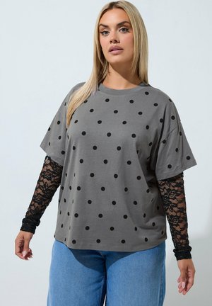 Vrouw draagt een grijs shirt met korte mouwen en polkadots over zwarte kanten lange mouwen en blauwe jeans, staand tegen een effen achtergrond.