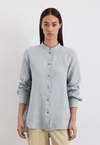 Chemise boutonnée gris clair avec col mandarin, manches longues et coupe décontractée. Elle présente une texture lisse et un ourlet arrondi.