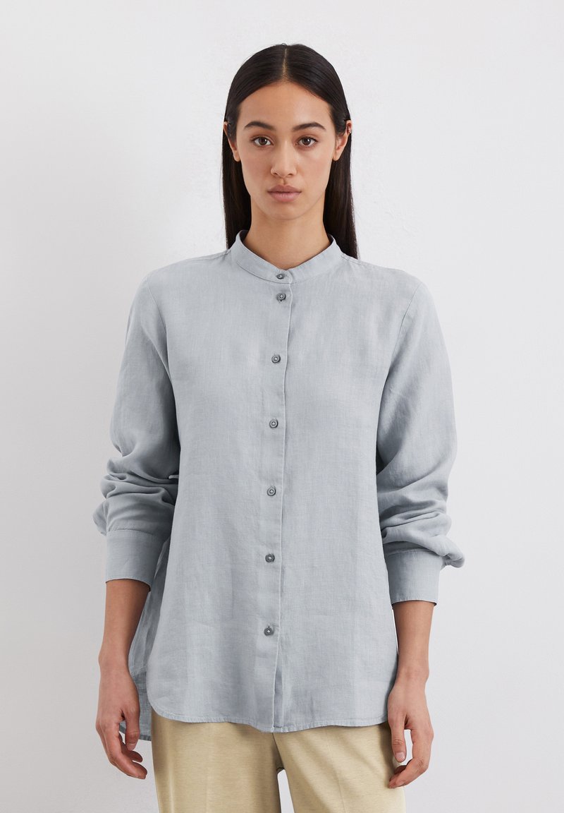 Chemise boutonnée gris clair avec col mandarin, manches longues et coupe décontractée. Elle présente une texture lisse et un ourlet arrondi.