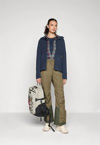 Mornarska flis jakna u kombinaciji s kariranom košuljom, maslinasto zelene hlače s crnim detaljima i krem cipele. Uključena je svijetlosiva ruksak.