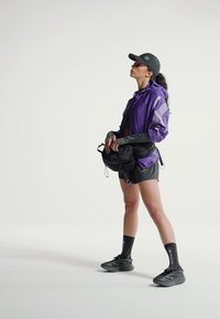 Donna vestita con giacca viola, pantaloncini neri, cappello nero e scarpe da ginnastica nere, che tiene una cintura nera portamonete, in piedi di profilo su uno sfondo bianco.