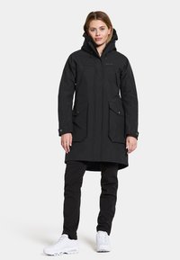 Parka nera impermeabile con cappuccio, dotata di due ampi tasconi frontali, taglio dritto e cuciture rinforzate. Indossata sopra pantaloni neri e scarpe bianche.
