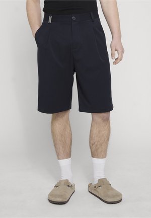 Male wearing black knee-length shorts, white socks, and beige slip-on shoes, standing with one hand in pocket and one relaxed by side.