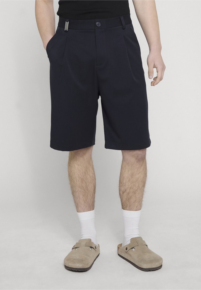 Mann trägt schwarze knielange Shorts, weiße Socken und beige Slipper, steht mit einer Hand in der Tasche und einer entspannten am Körper.