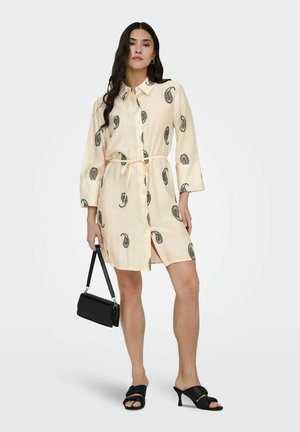 Kvinne iført en kremhvit skjortekjole med svarte paisleymønstre, holder en svart håndveske og har på seg svarte sandaler med hæl.