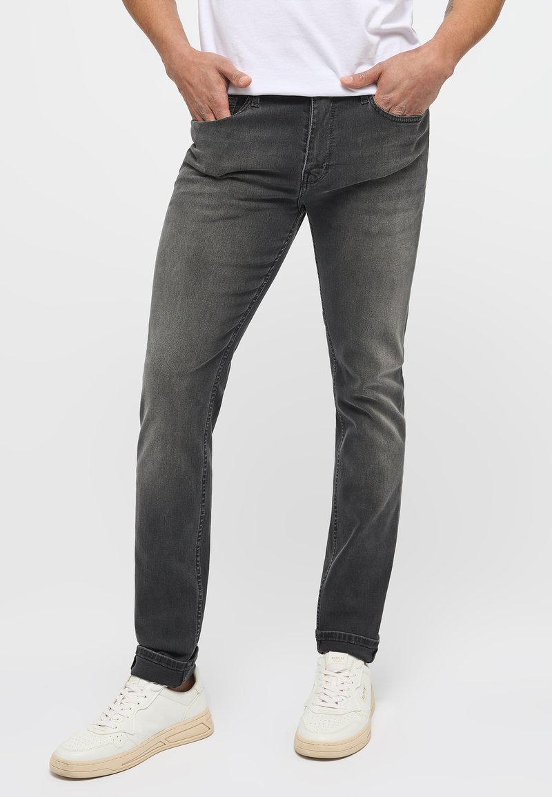 Szürke farmernadrág, slim fit szabással, egyenes lábszárral, öt zsebbe és diszkrét kopássa. Fehér sportcipővel és egyszerű fehér pólóval párosítva.