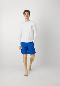 Vit långärmad rash guard med logotyp på bröstet, kombinerad med enfärgade blå badshorts. Modellen är barfota och står mot en neutral bakgrund.