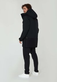 Svart vattentät parka med en stor huva, designad med en längre bakdel. Kombinationen med svarta byxor och vita sneakers ger en neutral kontrast.