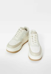 Bílé kožené tenisky s hladkým povrchem, perforovanou špičkou, béžovými akcenty a hnědou gumovou podrážkou. Součástí jsou tkaničky a polstrovaný límec.