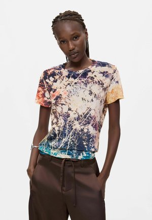 Donna con capelli intrecciati che indossa una t-shirt a maniche corte con stampa astratta multicolore e pantaloni marrone scuro con le mani in tasca.