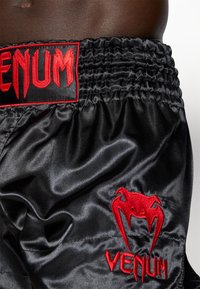 Calções de seda pretos com um cós franzido, com o texto bordado em vermelho "VENUM" e um logo de aranha. Textura suave e cores ousadas.