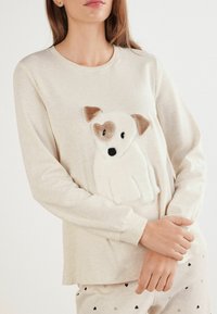 T-shirt beige à manches longues avec une appliqué en forme de chien blanc pelucheux et des accents marron. Tissu doux, col rond et coupe décontractée.