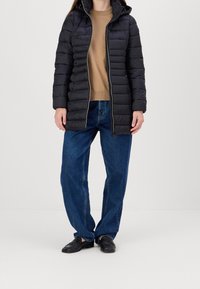Manteau doudoune noir avec capuche, design matelassé, fermeture éclair sur le devant et poches latérales, porté sur un pull beige et un jean large bleu.