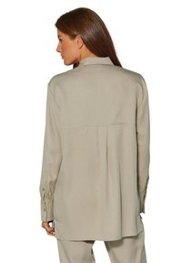 Beige Langarmshirt mit einer Rückenfalte, geknöpften Manschetten und einem abgerundeten Saum. Glattes, leichtes Material mit minimalen Nähdetails.