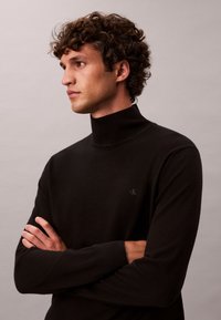 Joven con pelo rizado que lleva un suéter negro de cuello alto, brazos cruzados, mirando hacia la izquierda sobre un fondo liso.