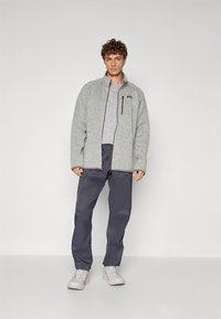 Γκρι φλις μπουφάν με φερμουάρ μπροστά, γκρι κοντομάνικο μπλουζάκι από κάτω, σκούρο μπλε παντελόνι και λευκά chunky αθλητικά παπούτσια. Διαθέτει τσέπη με φερμουάρ στο μπουφάν.