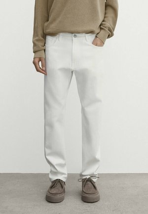 Osoba ubrana w białe spodnie o prostych nogawkach, beżowy sweter i szare zamszowe buty sznurowane, stojąca na szarej podłodze przy białej ścianie.