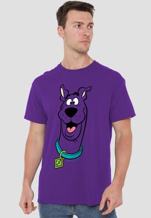 Mann trägt ein lila T-Shirt mit einer großen Cartoon-Grafik des lächelnden Gesichts von Scooby-Doo und blauem Halsband auf der Vorderseite.
