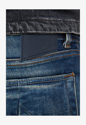 Jeans di denim con una toppa in pelle blu scuro con testo in rilievo. Cuciture arancioni e design delle tasche visibile in un lavaggio medio.