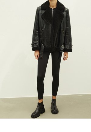 Veste en cuir noire avec une doublure en polaire, poches zippées et accents à boucles, portée avec des leggings noirs et des bottines brillantes.