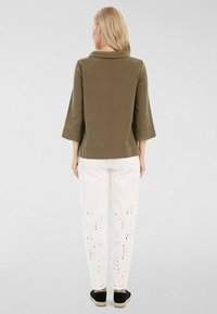 Veste vert olive à manches trois-quarts, texture lisse, associée à un pantalon blanc orné de détails en forme de feuilles découpées et à des espadrilles noires.